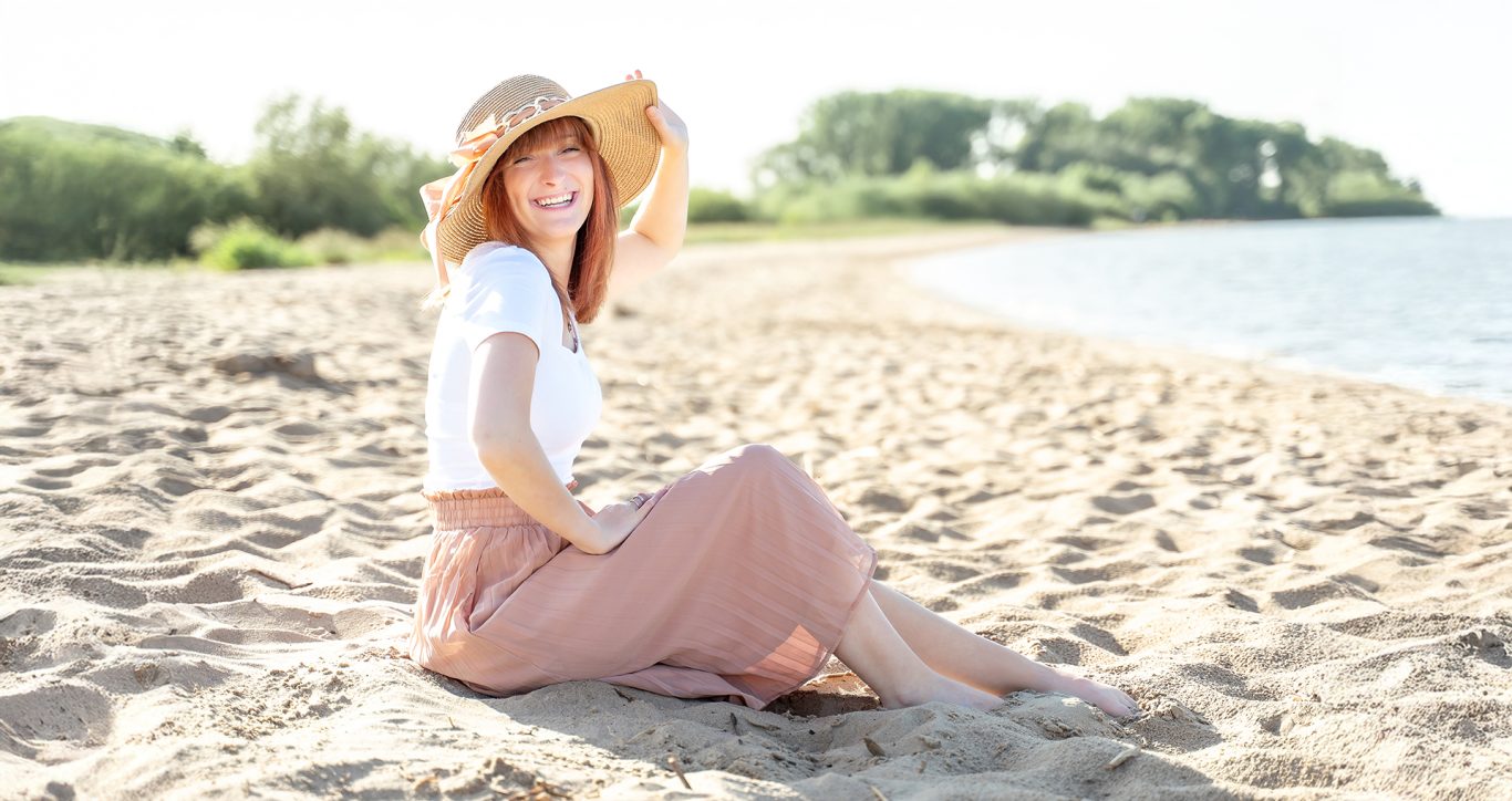 Frau mit Hut sitzt am Strand, sonnige Atmosphäre, lächelnd.